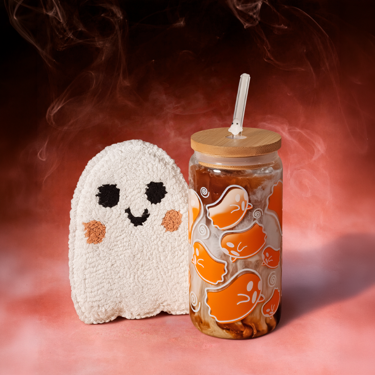 Orange ghost glass tumbler with a bamboo lid and ghost-charm straw beside a tufted ghost-shaped coaster against a smoky orange background.