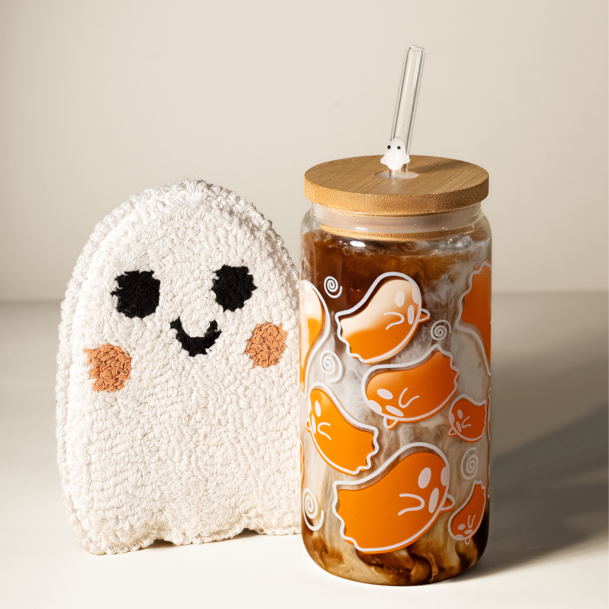 Glass tumbler with orange ghost designs, a bamboo lid and ghost-charm glass straw beside a white tufted ghost-shaped coaster.