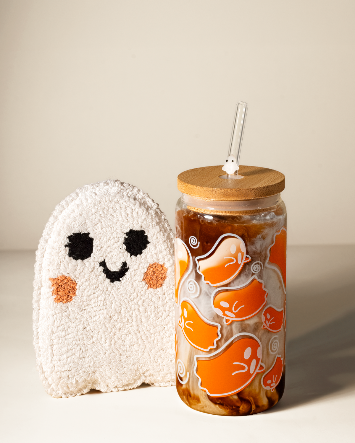 Orange ghost glass tumbler filled with an iced drink, with a bamboo lid, ghost-charm straw and tufted ghost-shaped coaster on a beige background.