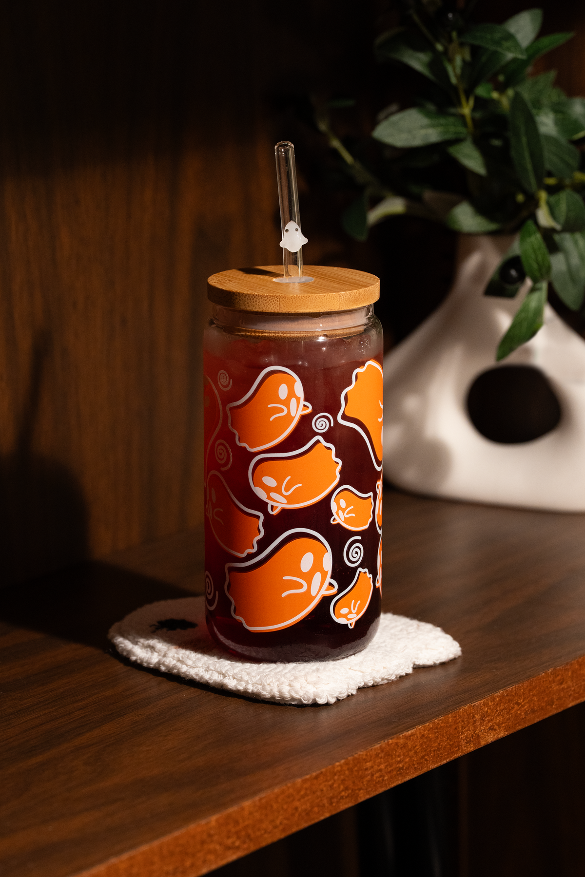 Orange ghost glass tumbler with a bamboo lid and ghost-charm straw on a white tufted coaster on a wooden shelf.
